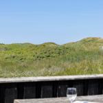 Terrasse mit Tisch und Gläsern, Blick auf grüne Dünen und klaren Himmel.