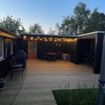 Holzterrasse mit Tisch, Stühlen und Grill. Hintergrund: Wald und Lichterkette.