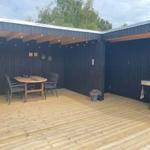 Terrasse mit Holzdecke, Tisch, Stühlen und Grill. Hintergrund: dunkle Holzwände und Bäume.