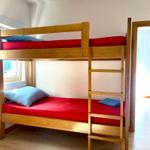 Zimmer mit Holzbett und roten Matratzen. Fenster und Tür sichtbar.