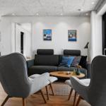Wohnzimmer mit grauem Sofa, zwei Sesseln und Holztisch. Große Fenster und Türen.
