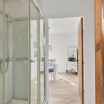 Shower area with glass enclosure. View into living area with table and chair.