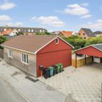 Red house with garage and paved yard. Several trash bins are in front of the building.