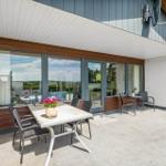 Terrasse mit Tisch, Stühlen und Blumen. Große Fensterfront zum Innenraum.