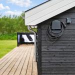 Außenansicht eines Holzhauses mit Ladekabel an der Wand und Terrasse mit Liegestühlen.