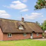 Rotes Backsteinhaus mit strohgedecktem Dach und weißen Fenstern in grüner Umgebung.