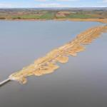 Schlankes Landstück mit Brücke führt über Wasser zu Land. Hintergrund: Felder und Horizont.