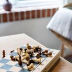 Schachbrett mit Figuren auf Tisch, Fenster und Stuhl im Hintergrund.