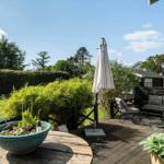 Terrasse mit Sitzgruppe, Sonnenschirm und Pflanzen. Grünfläche und Bäume im Hintergrund.