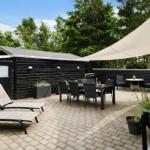 Terrasse mit Sonnensegel, Liegestühlen und Esstisch. Hintergrund: schwarzes Holzhaus und Bäume.