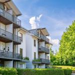 Wohnhaus mit Balkonen und grüner Umgebung unter blauem Himmel.