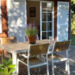 Terrasse mit Tisch und Stühlen, Pflanze und Glasflasche vor Haus mit Fenster und Jalousien.