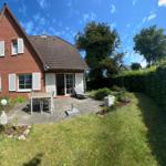 Rotes Backsteingebäude mit Terrasse, Garten und Sitzgelegenheiten unter blauem Himmel.