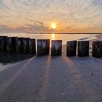 Sonnenuntergang am Strand mit Holzpfählen und Steg im Hintergrund.