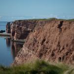 Rote Klippen am Meer mit Gras und Blick aufs Wasser.