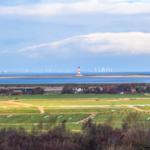 Weitläufige Landschaft mit Leuchtturm, Windkraftanlagen und grünen Feldern am Wasser.