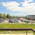 Blick auf Parkplatz, Grünfläche und Strandpromenade mit Gebäuden und Fahnen.