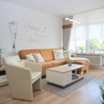 Wohnzimmer mit orange Sofa, weißen Sessel und Holzboden. Große Fenster mit Blick nach draußen.