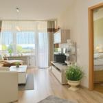 Wohnzimmer mit Sofa, Couchtisch und Fernseher. Blick auf Balkon. Schlafzimmer mit Bett sichtbar.