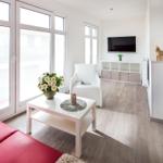 Wohnbereich mit rotem Sofa, weißem Sessel und TV. Große Fenster und Holzboden.