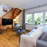 Wohnzimmer mit Sofa, Sessel und TV unter Holztreppe. Fenster mit Blick auf Bäume.