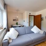 Ein Wohnzimmer mit grauem Sofa, weißen Kissen und Holzboden. An der Wand hängt eine Uhr.