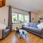 Wohnzimmer mit Sofa, Sessel und Holzboden. Fenster mit Blick auf Grün.