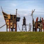 Pirate ship playground with slide and ladder on green slope. Background: black lighthouse.