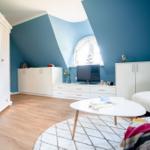 Zimmer mit blauen Wänden, weißer Einrichtung und Holzboden. Fenster und TV sichtbar.