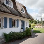 Haus mit Strohdach, blauen Fensterläden und blauer Tür. Garten mit Blumen und Rasen.
