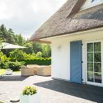 Haus mit Strohdach, Terrasse, Gartenmöbel und grüner Umgebung.