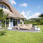 Haus mit Strohdach, Terrasse mit Liege und Sonnenschirm, umgeben von Gras und Bäumen.