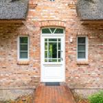 Haus mit rotem Ziegelstein, weißer Tür und strohgedecktem Dach.