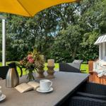 Tisch mit Kaffeetassen, Blumen und Buch unter gelbem Sonnenschirm. Strandkorb und Garten im Hintergrund.