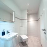 Badezimmer mit Dusche, Waschbecken und Toilette. Fliesen und Spiegel mit Beleuchtung.