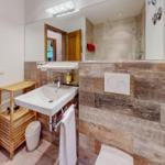 Badezimmer mit Waschbecken, Spiegel und Dusche. Holzwand und Toilette sind sichtbar.