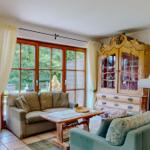 Wohnzimmer mit Sofa, Couchtisch und antikem Schrank. Große Fenster mit Blick auf Garten.