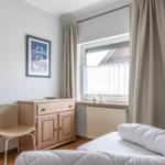 Schlafzimmer mit Bett, Holzschrank und Stuhl. Fenster mit Vorhängen und Bildern an der Wand.