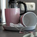 Auf der Arbeitsfläche stehen zwei Tassen, eine davon umgekippt, mit Teebeuteln und einem rosa Behälter.