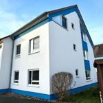 Zweistöckiges Haus mit weißen Wänden und blauen Akzenten. Viele Fenster und eine Eingangstür.