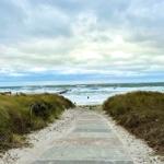Pfad durch Dünen zur Strandpromenade mit Leuchtturm im Hintergrund.