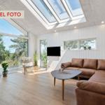 Wohnzimmer mit braunem Sofa, Holzboden und großen Fenstern. An der Wand hängt ein Fernseher.