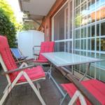 Terrasse mit Tisch und drei Stühlen mit roten Kissen. Hintergrund: Glasfront und grüne Hecke.