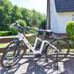 Ein weißes E-Bike steht vor einem Haus mit Garten und Hecke.