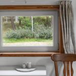 Ein Fenster mit Holzrahmen zeigt einen Blick in einen bewaldeten Bereich.