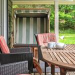 Terrasse mit Holztisch, Wickerstühlen und Strandkorb. Grün hinter dem Haus.