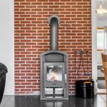 Ein Kaminofen steht vor einer Backsteinwand. Links ein Bücherregal, rechts ein Holztisch mit Stühlen.