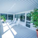 Terrasse mit Liegestühlen, Pflanzen und Glaswänden. Fliesenboden und weißer Dachbalken.