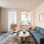 Wohnzimmer mit Sofa, Sessel und Tisch. Küche im Hintergrund sichtbar.