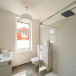 Bad mit Dusche, Waschbecken und Toilette. Fenster mit Blick auf rotes Gebäude.